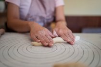 Обрізати безлику жінку в фартухах, що прокатує тісто руками на столі, готуючи домашні пельмені на кухні — стокове фото