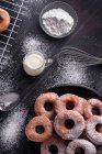 Сладкие жареные пончики подаются на тарелке возле металлической стойки охлаждения и кувшин молока на черном грязном столе с сахарной пудрой — стоковое фото