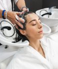 Crop unrecognizable master washing dark hair of female customer with closed eyes in beauty salon — Stock Photo