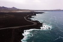 Drohnenblick auf schäumendes Meer gegen gebogene Fahrbahn und Berge mit Hervideros in Yaiza Lanzarote Kanarische Inseln Spanien — Stockfoto