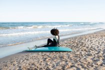 Back view full body of anonymous barefoot female surfer in wetsuit sitting on sandy beach near waving sea — Stock Photo