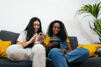 Jovens amigas diversas em roupas casuais sorrindo enquanto se sentam no sofá navegando no smartphone na sala de estar em casa — Fotografia de Stock