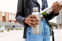 Anonyme afroamerikanische Studentin mit Thermoskanne und ein paar Schulbüchern stehen auf der Straße in der Nähe des Universitätsgebäudes — Stockfoto