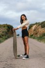 Full body side view of young smiling ethnic cheerful female standing with longboard and headphones on neck looking at camera between hills under cloudy sky — Stock Photo