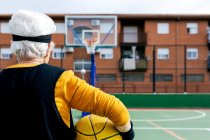 Анонимный спортсмен в спортивной одежде, стоящий на площадке с желтым мячом и баскетбольным кольцом во время игры на улице — стоковое фото