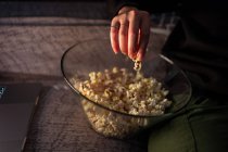 Zugeschnittene, unkenntlich gemachte Frau in lässiger Kleidung sitzt mit überkreuzten Beinen auf der Couch und isst zu Hause Popcorn — Stockfoto