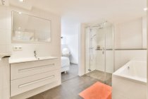 Современная белая ванная комната с душевой кабиной и ванной возле раковины под зеркалом в современной квартире — стоковое фото