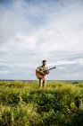 Спокойный человек музыкант в повседневной одежде, стоящий среди зеленой травы на берегу океана и играющий на акустической гитаре летом при дневном свете — стоковое фото