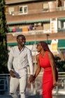 Alegre pareja afroamericana en ropa de moda tomados de la mano y riendo mientras caminan juntos en la calle de la ciudad en un día soleado - foto de stock