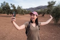Deliziosa viaggiatrice asiatica a braccia aperte che si autoritrae sullo smartphone mentre si trova in piantagione con ulivi in campagna — Foto stock