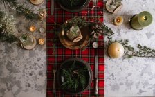 Вид на сервировку рождественского стола с венком на тарелке, декоративные деревянные украшения и красную клетчатую скатерть с желтыми огнями на заднем плане — стоковое фото