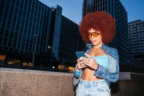 Mulher atraente com penteado afro e roupas na moda mensagens de texto no celular enquanto está de pé na rua com edifícios modernos à noite — Fotografia de Stock