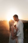 Homem abraçando mulher macia de pé perto entre cavalos calmos no campo montanhoso na luz do pôr do sol com os olhos fechados — Fotografia de Stock