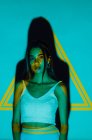 Cool young ethnic female in crop top looking at camera against yellow triangle and shadow — Stock Photo