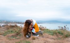 Heureux voyageur femelle étreignant chiot mignon tout en étant assis avec les jambes croisées sur la prairie dans les hauts plateaux à playa de las catedrales en Galice au coucher du soleil — Photo de stock