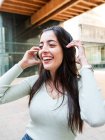 Fröhliche Frau mit langen braunen Haaren in lässiger Kleidung und Kopfhörern, die Musik hört und bei Tageslicht auf der Straße lacht — Stockfoto