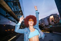 Happy female with Afro hairstyle in stylish outfit standing with raised arms on street with residential buildings in evening time — Stock Photo