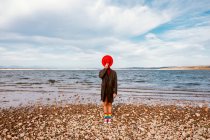 Persona irreconocible en sombrero rojo y abrigo de pie cerca de la orilla con piedras trituradas en verano - foto de stock