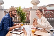 Side view of positive female in wireless earbuds showing cellphone to male flatmate working on laptop at wooden table on balcony — Stock Photo