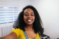Blogueiro feminino afro-americano feliz com cabelos longos encaracolados em camiseta amarela e jeans em geral em pé com mão no bolso e tomando selfie no smartphone na sala de luz — Fotografia de Stock