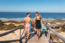 Back view full body of sportive couple with surfboards strolling together on wooden path near green plants before training in tropical resort — Stock Photo