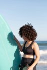 Vista laterale di felice giovane surfista donna in muta con tavola da surf in piedi guardando lontano sulla riva del mare lavata dal mare ondulato — Foto stock