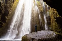Back view of unrecognizable tourist in warm hooded jacket standing in rocky canyon with powerful picturesque Gljufrafoss waterfall with outstretched arms during trip in Iceland — Stock Photo
