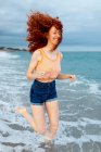 Full body of happy barefoot female traveler with flying long red hair running along sandy beach washed by foamy waves in windy weather — Stock Photo
