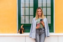 Positive Frau in stylischem Outfit mit Kaffee zum Mitnehmen schaut in die Kamera, während SMS auf dem Handy in der Nähe des Gebäudes mit Handtasche — Stockfoto