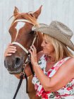 Нежная женщина-всадница держит уздечку каштановой лошади во дворе в летний день в сельской местности — стоковое фото