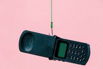 Old black opened flip mobile phone hanging on metal hook with green rope against pink background in light modern studio — Stock Photo