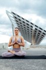 Full body of peaceful female in activewear with closed eyes practicing Padmasana posture on street against modern solar panel in city — Stock Photo