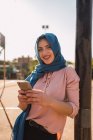 Mujer árabe joven positiva en hijab navegando por el teléfono móvil mientras está de pie cerca de polo en la calle de la ciudad y mirando a la cámara - foto de stock