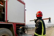 Полная длина храбрый анонимный пожарный в специальной защитной форме и держа топор на плече, стоя рядом с красной пожарной машиной и глядя в сельскую местность — стоковое фото