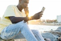 Crop African American male in casual clothes sitting with hand at chin on rocky coastline while using smartphone in summer evening — Stock Photo