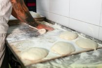 Vista lateral de la cosecha de panadero masculino tatuado en polo blanco amasando masa con las manos mientras está de pie en el mostrador de metal en la cocina de la panadería moderna - foto de stock