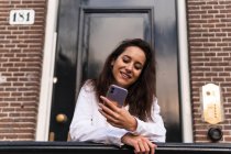 Fröhliche junge Frau in legerer Kleidung steht am Eingang des Gebäudes und lehnt am Geländer, während sie das Smartphone benutzt — Stockfoto