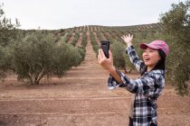 Délicieuse voyageuse asiatique avec les bras écartés prenant autoportrait sur smartphone tout en se tenant debout sur la plantation d'oliviers dans la campagne — Photo de stock