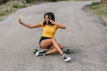 Full body of happy ethnic female sitting on longboard with crossed legs while showing peace gesture and taking self portrait on cellphone — Stock Photo
