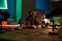Positif jeune homme barbu millénium en lunettes et écouteurs souriant et parlant en micro tout en enregistrant podcast en studio sombre — Photo de stock