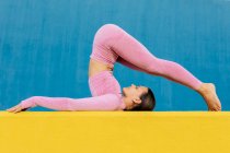 Vista lateral de la hembra delgada flexible en ropa deportiva haciendo ejercicio sobre alfombra amarilla contra la pared azul brillante - foto de stock