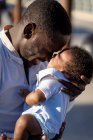Vista lateral de un cuidadoso padre afroamericano en camisa de pie y abrazando al pequeño bebé con el pelo rizado en la calle en un día soleado - foto de stock
