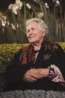 Vista lateral da mulher idosa pensativa com cabelos grisalhos embrulhando em xadrez sentado à mesa do jardim e olhando para longe no dia ensolarado — Fotografia de Stock