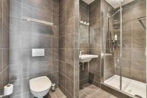 Белый туалет расположен у стены с коричневой плиткой рядом с раковиной с краном в светлом стильном туалете с душем и ванной — стоковое фото