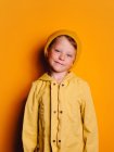 Счастливый мальчик в ярко-желтой куртке из дождевика и шапочке, смеющийся и смотрящий в камеру на жёлтом фоне в студии — стоковое фото