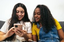 Jovens amigas diversas em roupas casuais sorrindo enquanto se sentam no sofá navegando no smartphone na sala de estar em casa — Fotografia de Stock