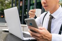 Beschnitten bis zur Unkenntlichkeit männliche ethnische Unternehmer mit Kaffee im Internet surfen auf dem Handy, während sie am städtischen Cafétisch mit Laptop sitzen — Stockfoto