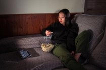 Молодая азиатка в повседневной одежде сидит на диване и ест попкорн дома, смотрит кино на ноутбуке — стоковое фото