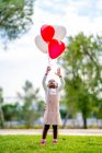 Веселая афро-американская девушка с косичками в стильной одежде, бегущая с красочными воздушными шарами в руке в парке днем — стоковое фото