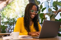 Positive African American female freelancer browsing internet on netbook while sitting at table with drink and notebook in outdoor cafeteria — Stock Photo